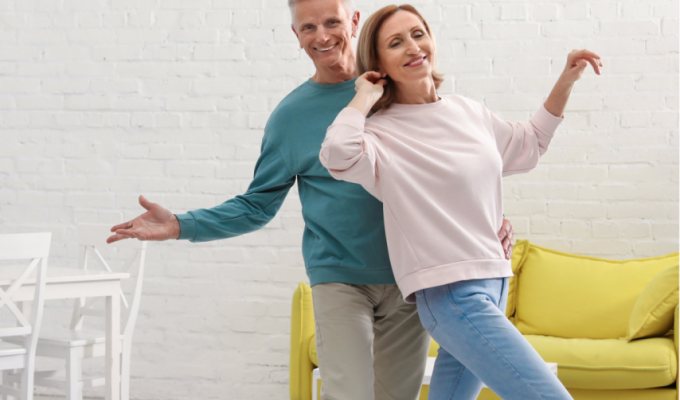 senior i seniorka tańczą w salonie na tle żółtej kanapy